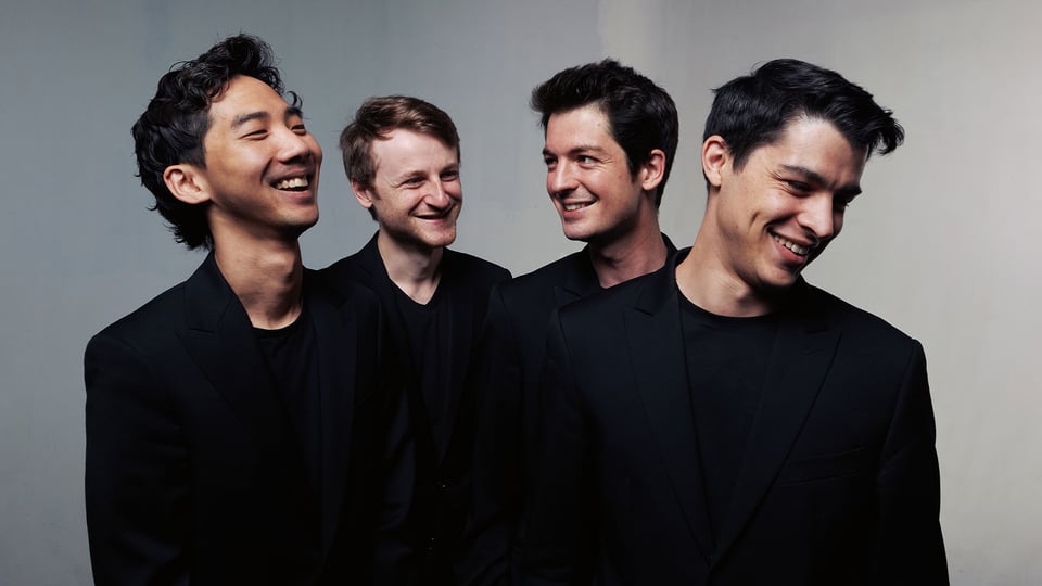 Quatuor Arod mit vier Musiker:innen in schwarzen Anzügen vor hellem Hintergrund, nebeneinander stehend und einander zugewandt, ohne Instrumente im Bild sichtbar.
