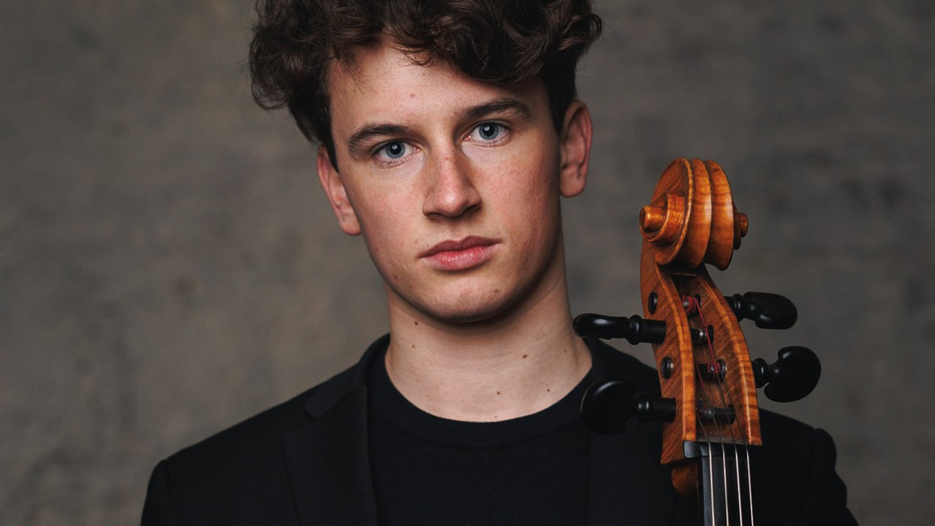 Der Cellist Lenz Defregger in schwarzem Pullover mit Instrument vor verschwommenem dunkelgrauem Hintergrund 