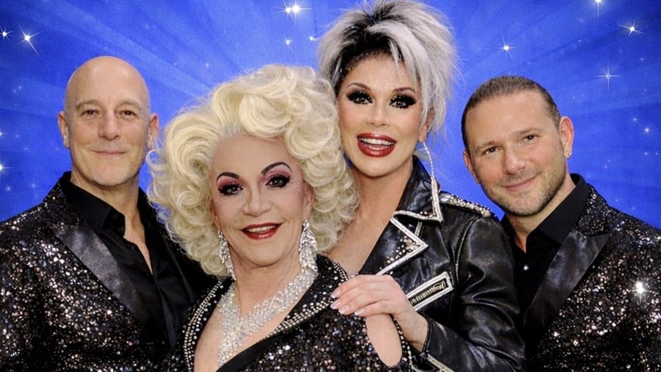 Vier Personen in glamourösen Bühnenoutfits posieren vor blauem, sternenartigem Hintergrund; im Zentrum zwei Travestiekünstlerinnen mit auffälligem Make-up und voluminösen Frisuren, flankiert von zwei Männern in glitzernden Anzügen. Promo-Bild zur Veranstaltung „25 Jahre Piggy & Gene – Travestierevueshow 2026“.