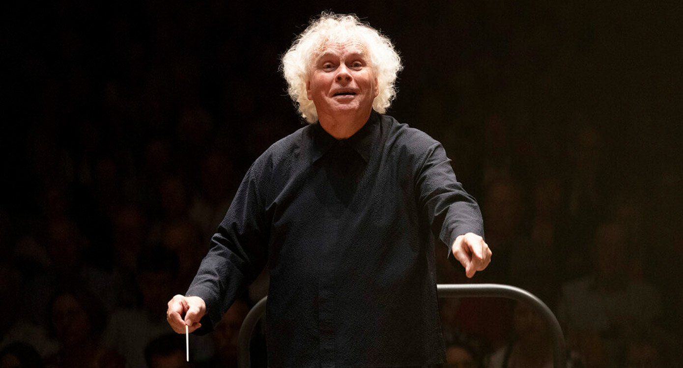 Dirigent Sir Simon Rattle in schwarzem Pullover mit Dirigentenstab vor dunklem Hintergrund
