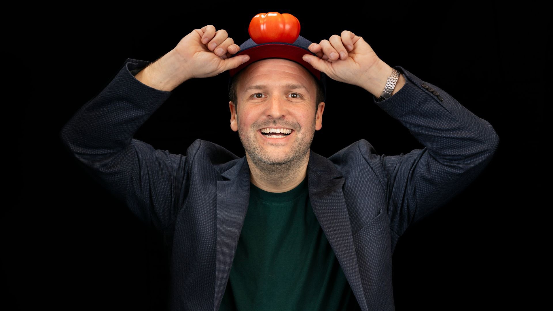 Christian Burmeder hält eine Mütze mit einer kleinen orangefarbenen Requisite darauf und hebt beide Hände an den Mützenrand, vor schwarzem Hintergrund