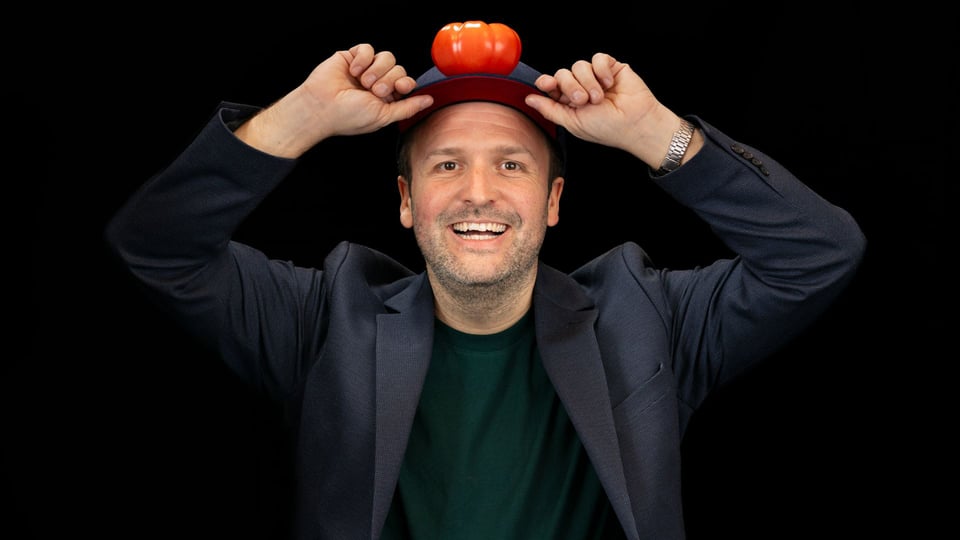 Christian Burmeder hält eine Mütze mit einer kleinen orangefarbenen Requisite darauf und hebt beide Hände an den Mützenrand, vor schwarzem Hintergrund