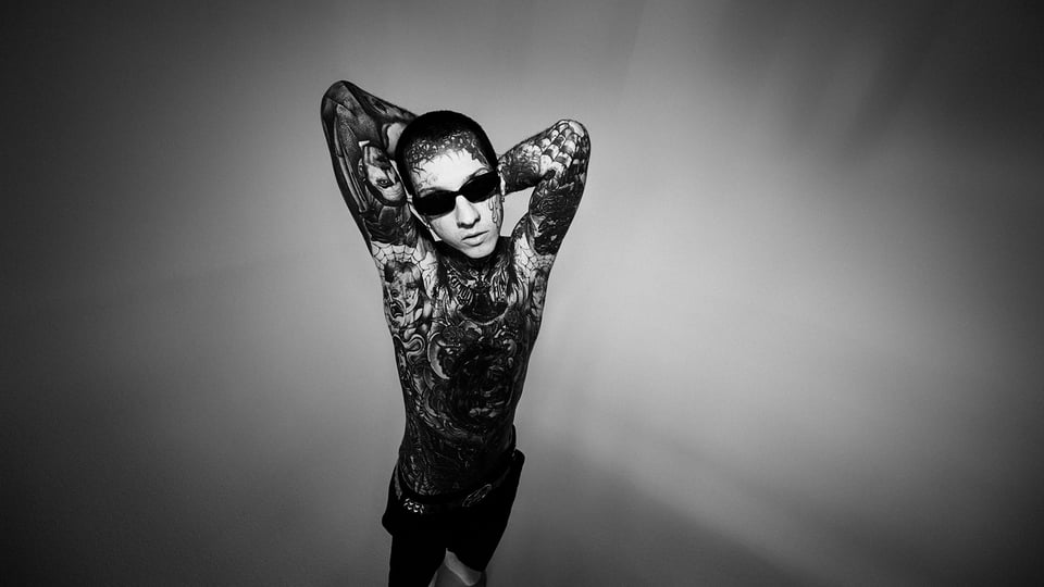 Gvllow in Schwarz-Weiß: tätowierter Künstler mit nacktem Oberkörper und Sonnenbrille steht vor neutralem Hintergrund, Arme hinter dem Kopf verschränkt, markanter, alternativer Look.