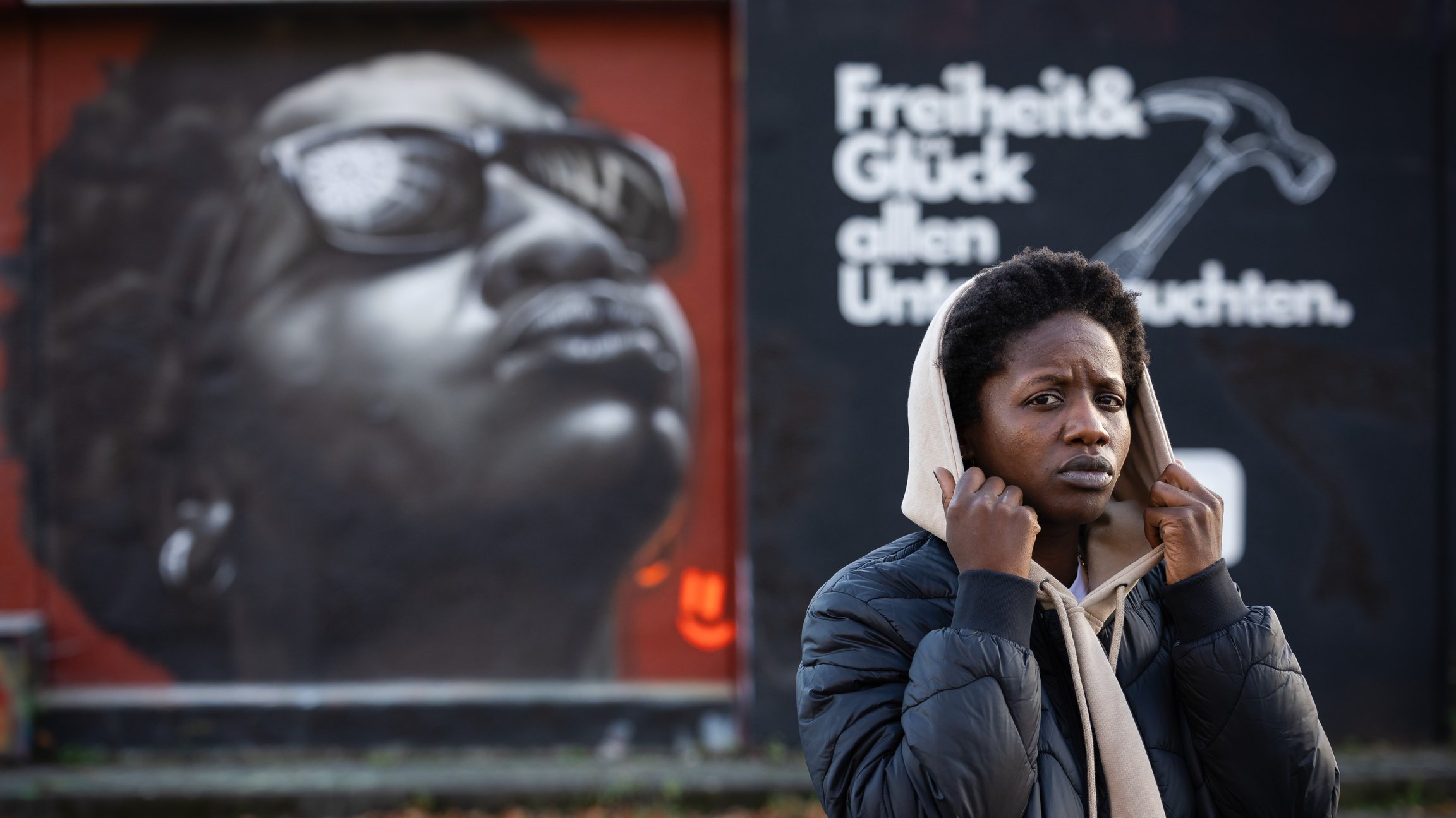 EIne Frau hält ihre Kapue fest, im Hintergrund ein Plakat mit einem Portrait und weißer Aufschrift