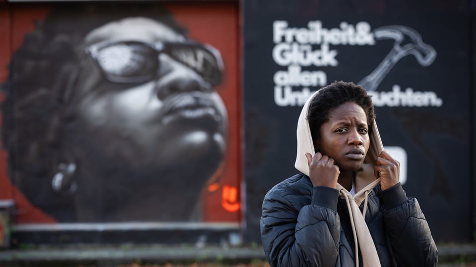 EIne Frau hält ihre Kapue fest, im Hintergrund ein Plakat mit einem Portrait und weißer Aufschrift