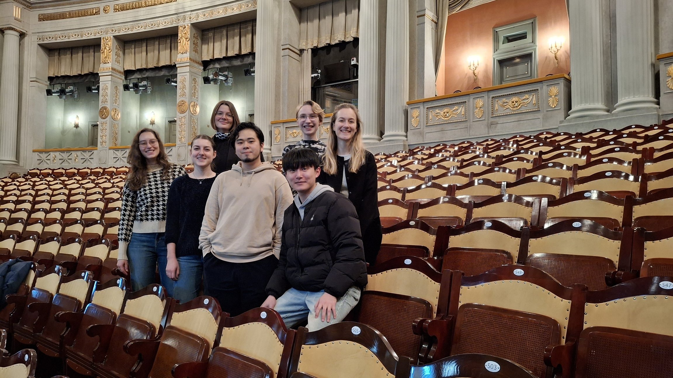 Gruppe von jungen Erwachsenen steht zwischen Reihen von Holzsitzen in einem historischen Theatersaal mit Säulen und dekoriertem Innenraum.