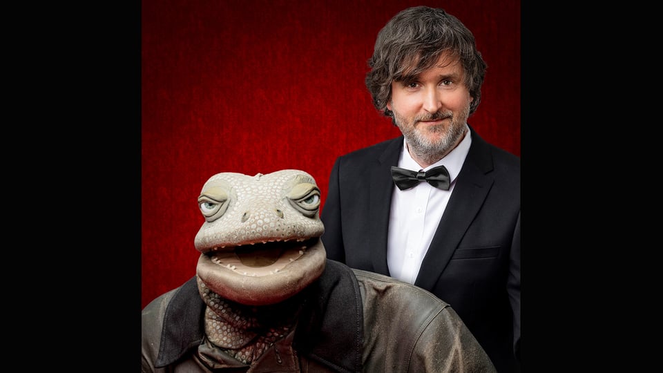 Porträtfoto zu „Michael Hatzius – ECHSKLUSIV“ mit Michael Hatzius im Anzug neben einer Echsenfigur vor rotem Hintergrund.