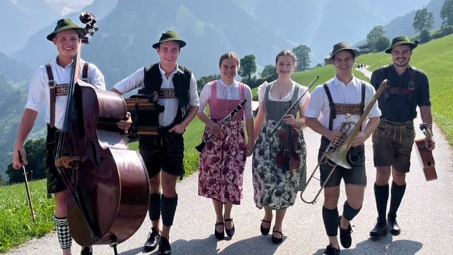 Sechs Personen in traditioneller Trachtenkleidung gehen gemeinsam auf einer Straße durch eine alpine Landschaft; sie tragen Instrumente wie Kontrabass, Trompete, Klarinette und Geige. Im Hintergrund sind grüne Wiesen, Berge und ein klarer Himmel zu sehen.