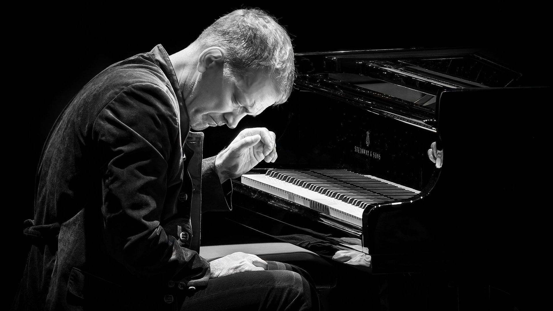 Schwarzweißes Foto von Brad Mehldau an einem Konzertflügel, seitlich auf einem Klavierhocker sitzend, mit gesenktem Kopf und erhobener Hand über der Klaviatur vor schwarzem Hintergrund