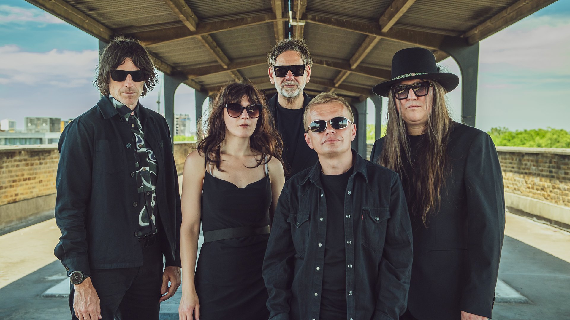 Bandfoto von Archive mit fünf Bandmitgliedern in schwarzer Kleidung und Sonnenbrillen unter einer überdachten Passage, darunter eine Person mit breitkrempigem Hut und eine Person in schwarzem Kleid, vor urbanem Hintergrund