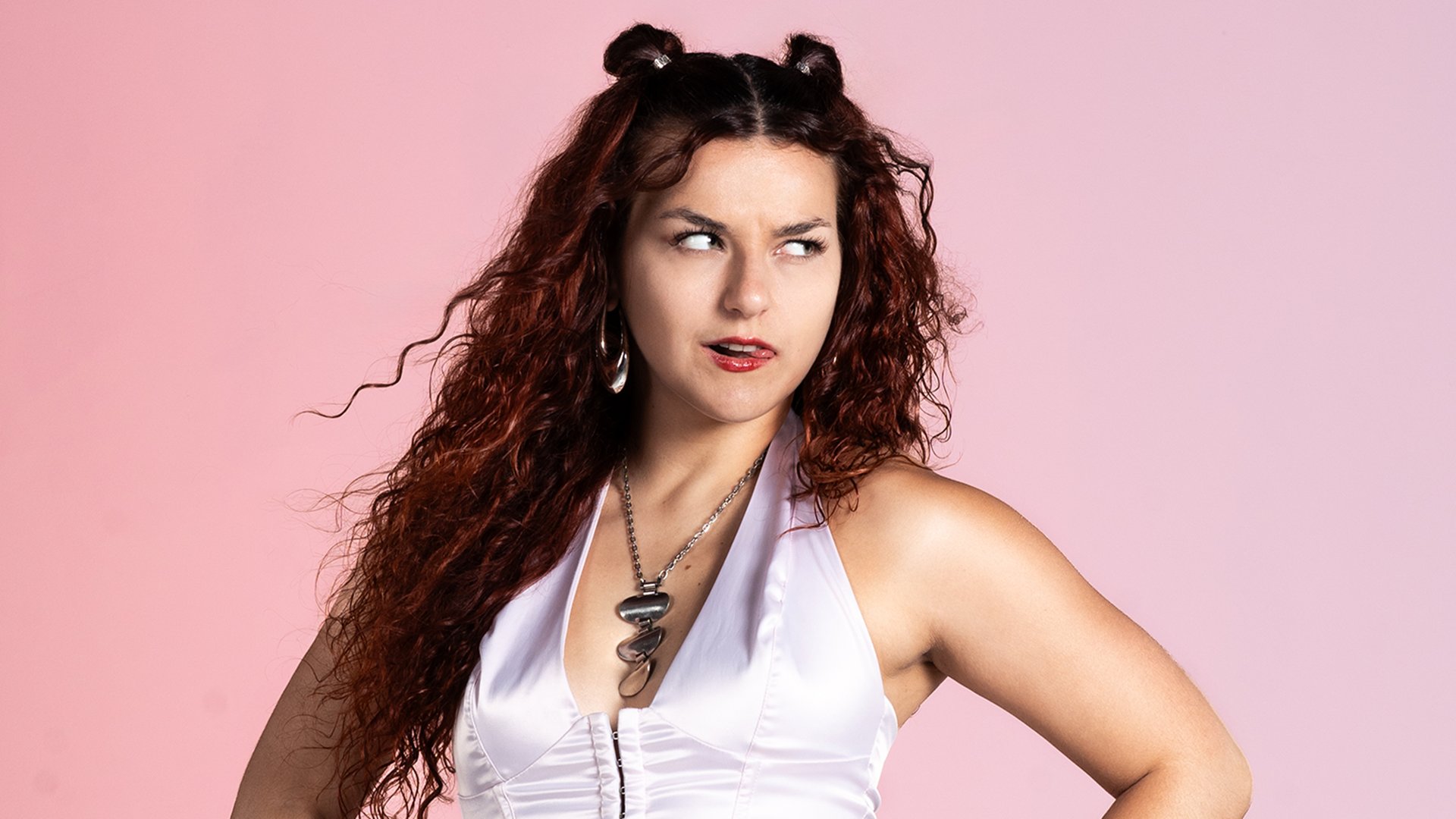 Portrait von Maiva vor hellrotem Hintergrund. Die Künstlerin schaut frech rechts neben der Kamera vorbei und streckt die Zunge raus.