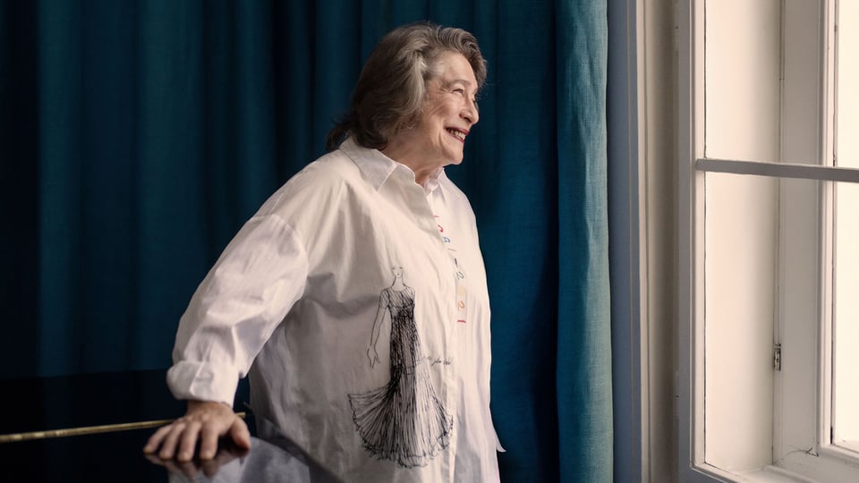 Elisabeth Leonskaja steht neben einem Fenster, trägt ein helles Hemd mit Zeichnung und blickt nach außen.