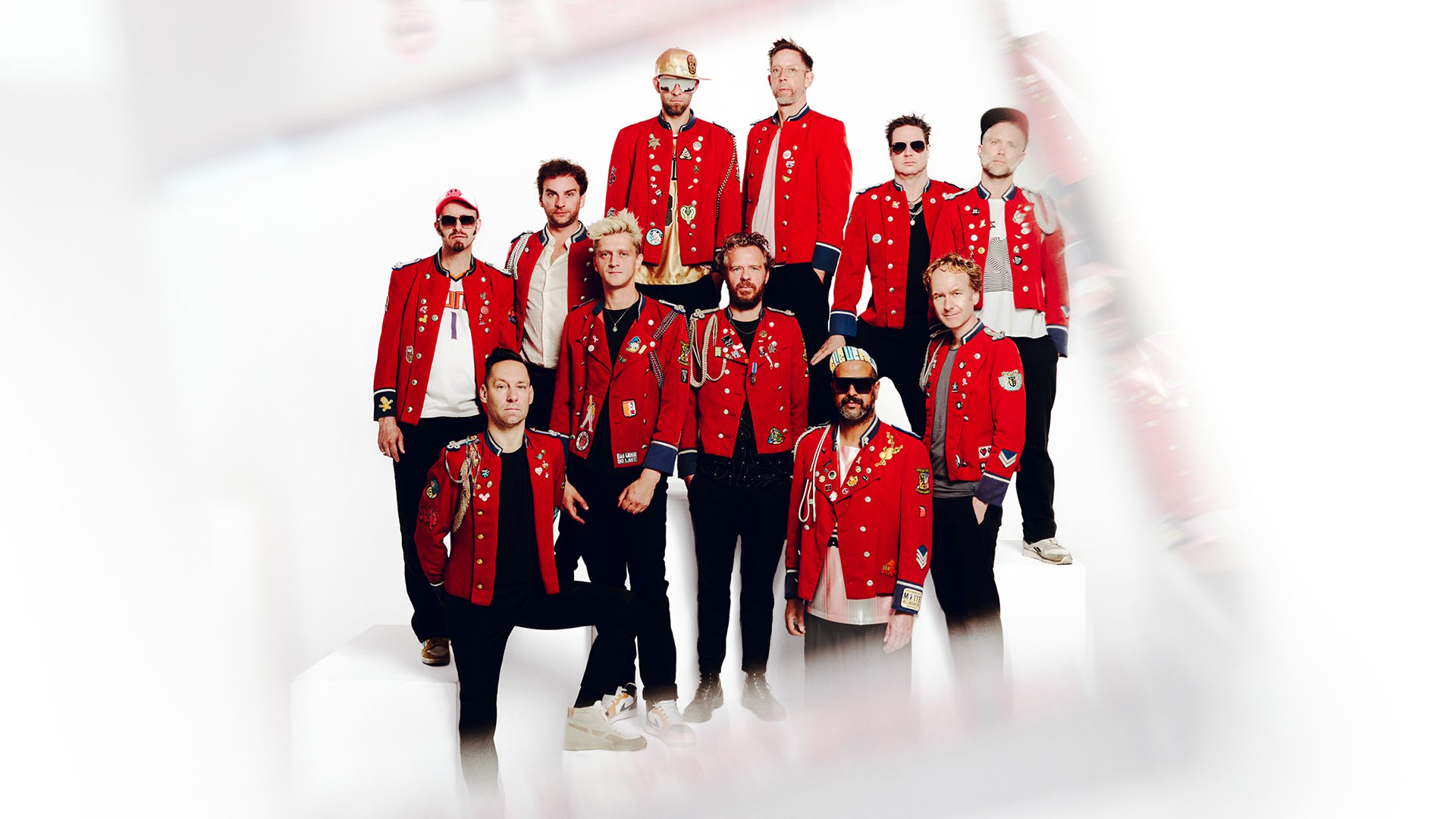 Band MEUTE mit mehreren Musikerinnen und Musikern in roten Uniformjacken steht auf weißen Podesten vor hellem Hintergrund und posiert als Gruppe.
