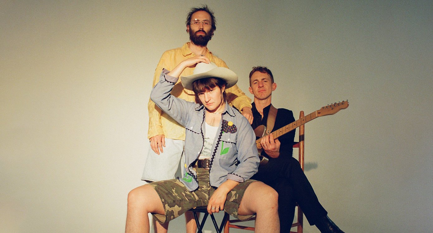 Drei Männer, einer im Vordergrund mit kurzer Lederhose und Hut, einer mit Gitarre
