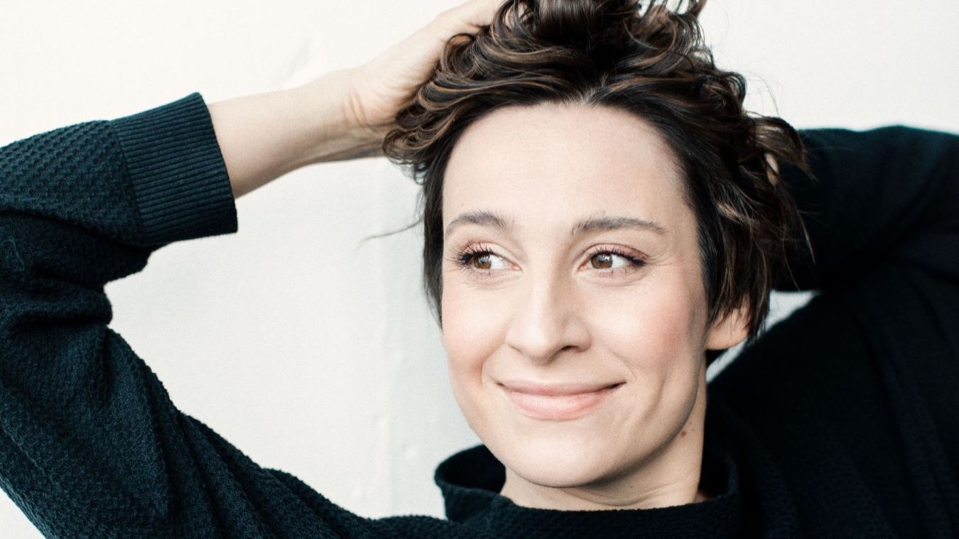 Porträt der Mezzosopranistin Christina Bock, die mit beiden Händen durch ihr kurzes Haar greift und leicht lächelt.