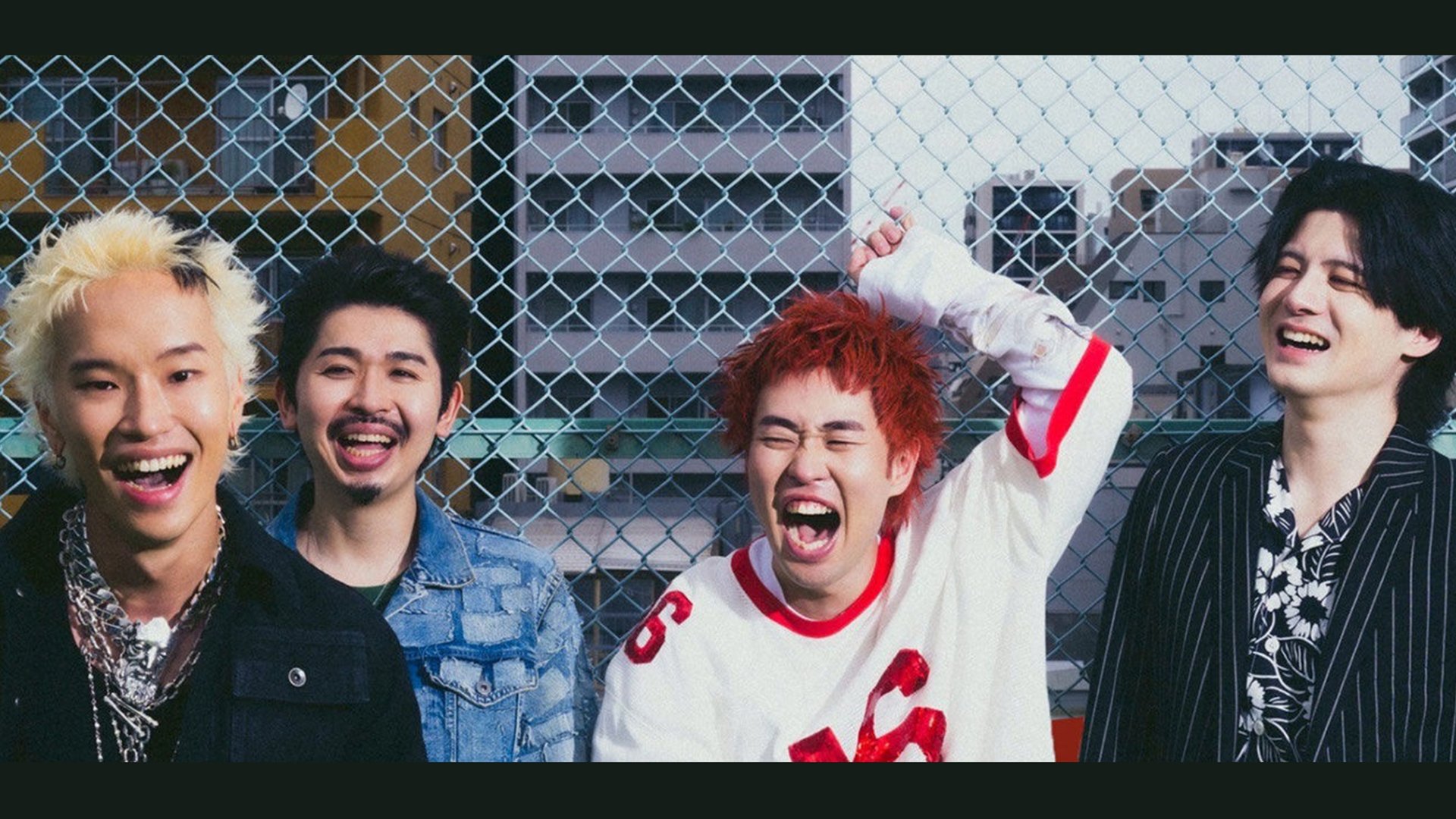 Die vier Bandmitglieder von Paledusk lachen vor einem Maschendrahtzaun auf einem urbanen Dach; bunte Haarfarben, lässige Kleidung und ausgelassene Stimmung, im Hintergrund Hochhäuser