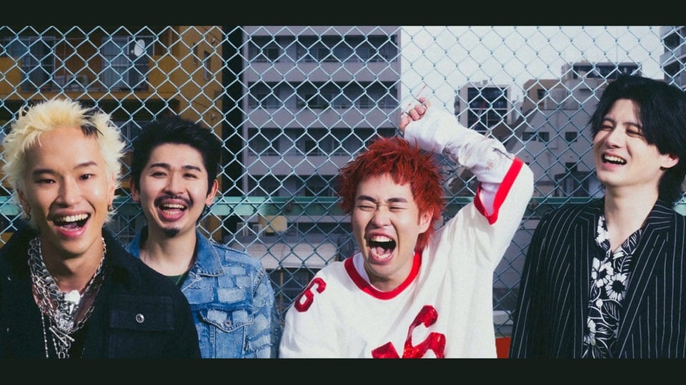 Die vier Bandmitglieder von Paledusk lachen vor einem Maschendrahtzaun auf einem urbanen Dach; bunte Haarfarben, lässige Kleidung und ausgelassene Stimmung, im Hintergrund Hochhäuser