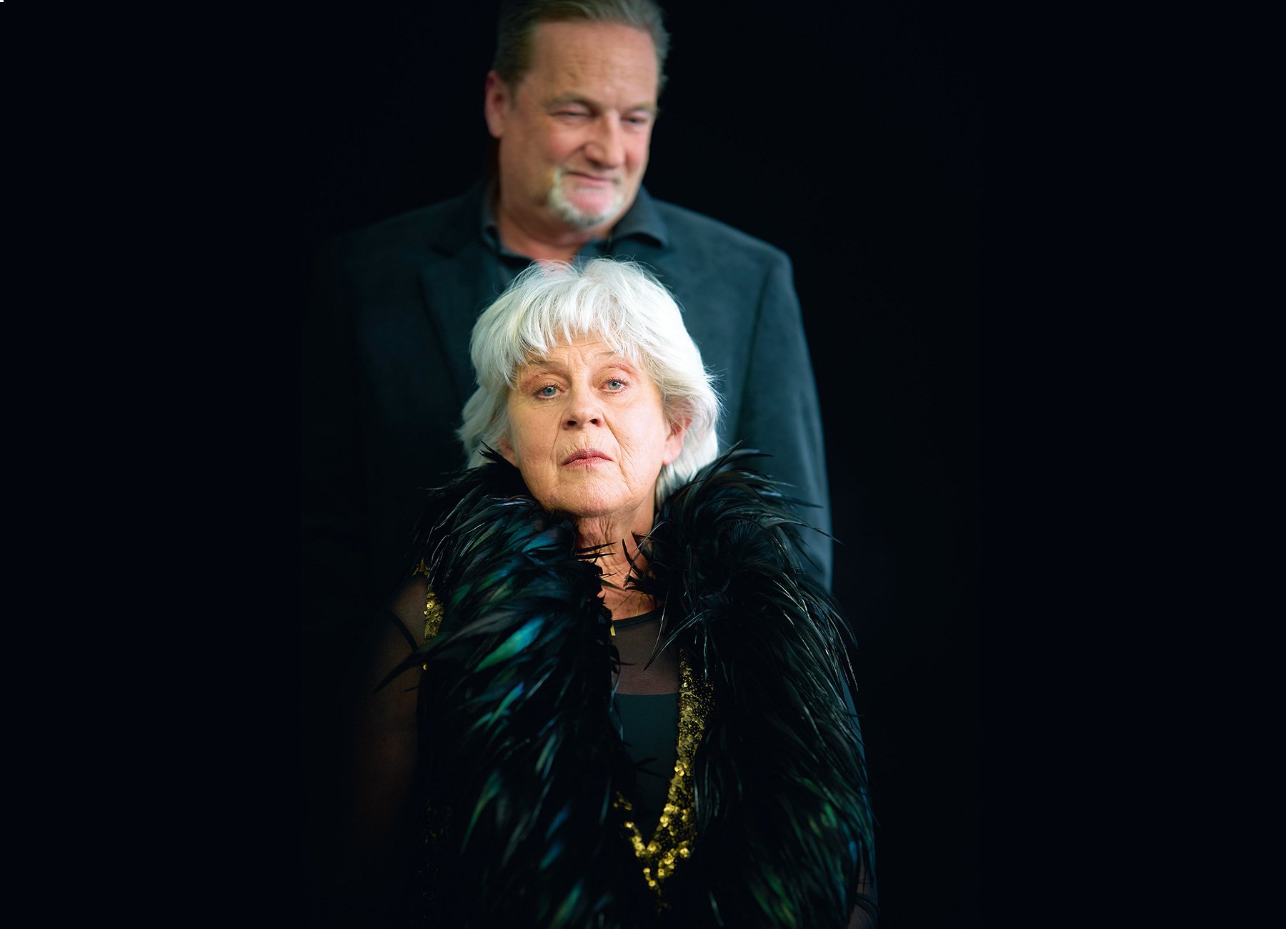 Eine Dame mit Federboa und ein Herr dahinter vor schwarzem Hintergrund