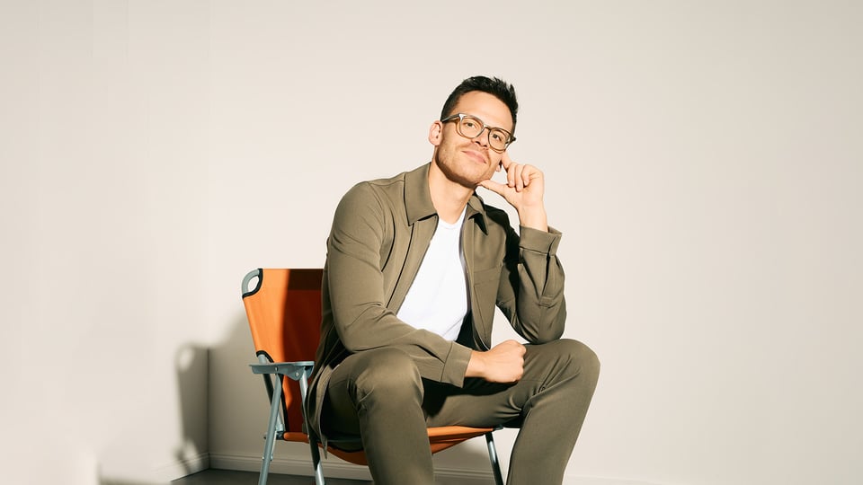 Martin Frank trägt ein grünes Outfit und sitzt vor einer hellen Wand auf einem orangenen Camping Stuhl.