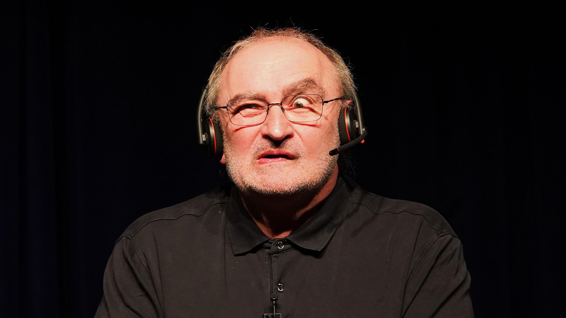 Portrait von Sigi Zimmerschied, der eine Grimasse macht, in schwarzem Hemd, mit Kopfhörern, vor schwarzem Hintergrund