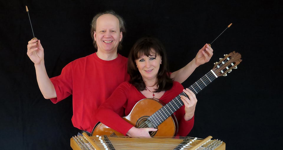 Ein Herr und eine Dame in roten Pullovern mit Instrumenten vor schwarzem Hintergrund