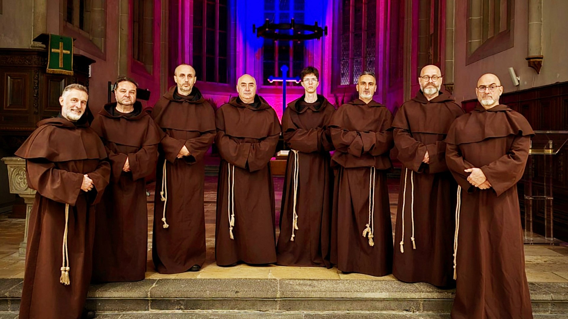 Acht Sänger in braunen Mönchskutten stehen in einer Kirche vor einem beleuchteten Kreuz und farbigem Bühnenlicht