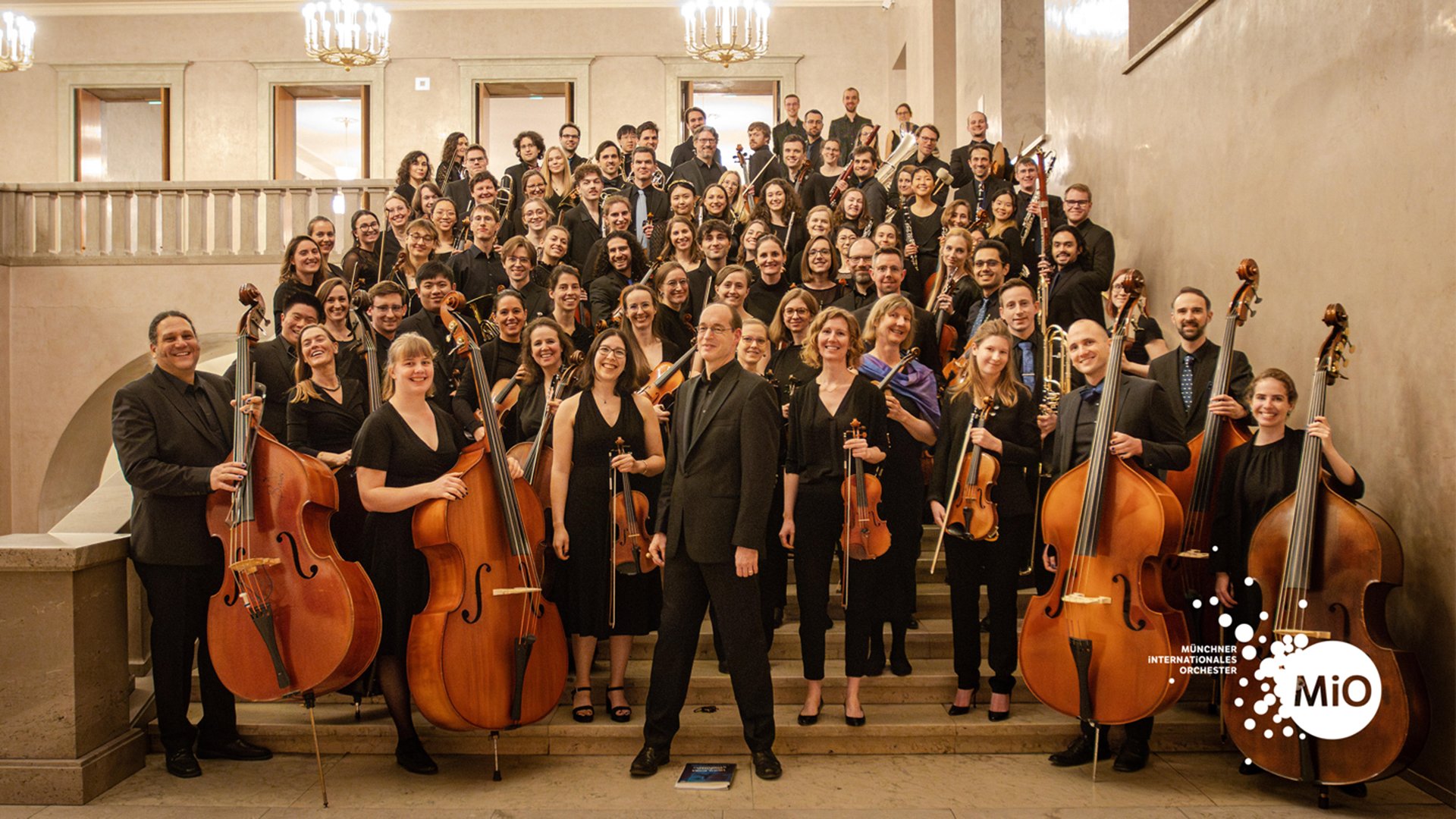 Gruppenfoto des Münchner Internationalen Orchesters (MiO) mit Musiker:innen und klassischen Instrumenten in schwarzer Konzertkleidung.