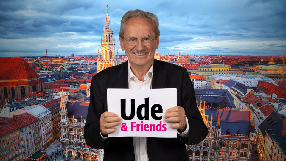 Person mit Brille hält ein Schild mit der Aufschrift Ude & Friends vor sich, im Hintergrund Panorama der Münchner Altstadt mit Rathaus, Kirchturm und Dächern bei Abendlicht