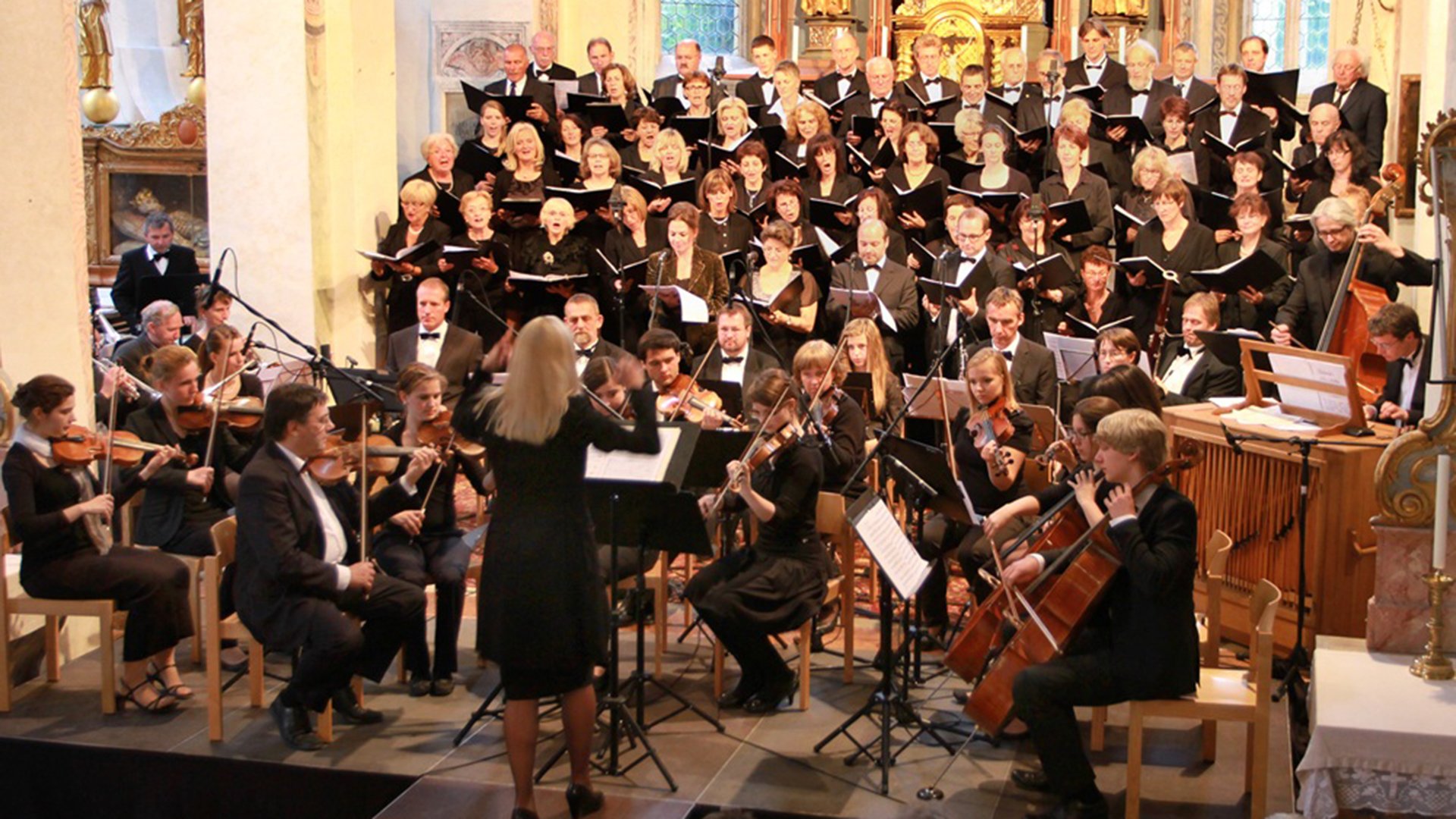 Festliches Pfingstkonzert im Mozart-Jubiläumsjahr mit Chor und Orchester in einer barocken Kirche, Dirigentin vor Ensemble mit Streichern und Sängerinnen und Sängern auf mehreren Ebenen vor Altarraum.