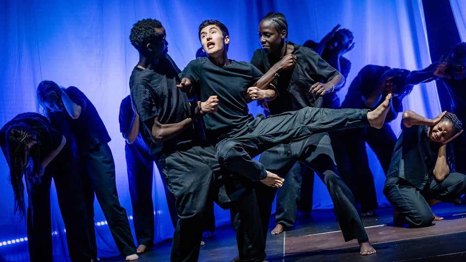 Bühnenfoto einer Tanztheatergruppe in dunkler Kleidung auf blau beleuchteter Bühne, zwei Personen tragen eine dritte Person im Mittelpunkt der Szene, während weitere Mitwirkende in verschiedenen Körperhaltungen im Hintergrund agieren