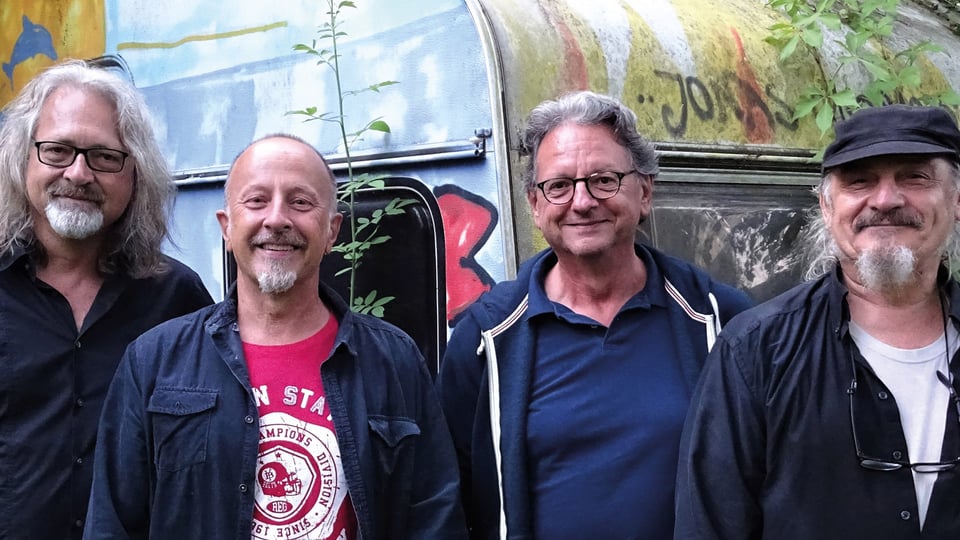 Vier Personen stehen nebeneinander vor einem alten, teilweise mit Graffiti besprühten Wohnwagen im Freien. Die Personen tragen überwiegend dunkle Freizeitkleidung, eine Person trägt ein rotes T-Shirt. Im Hintergrund sind Pflanzen und Bäume zu sehen, die dem Bild eine natürliche, rustikale Atmosphäre verleihen. Die Aufnahme zeigt die Gruppe in einer lockeren, informellen Umgebung.
