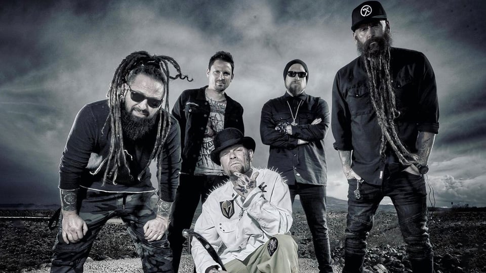 Die Band Five Finger Death Punch in dunkler Kleidung steht im Freien, markanter Himmel im Hintergrund, eine Person mit Hut im Vordergrund