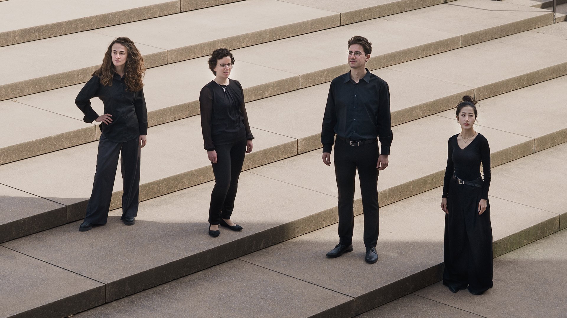 Vier Personen in schwarzer Kleidung stehen mit Abstand zueinander auf breiten Steinstufen im Außenbereich; die Szene wirkt geordnet und minimalistisch, mit klaren Linien und neutralem Hintergrund