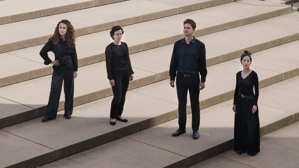 Vier Personen in schwarzer Kleidung stehen mit Abstand zueinander auf breiten Steinstufen im Außenbereich; die Szene wirkt geordnet und minimalistisch, mit klaren Linien und neutralem Hintergrund
