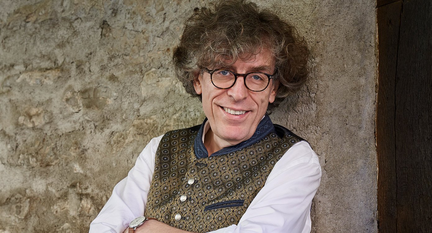 Mann mit Locken und Brille  in Brokatweste vor Mauer lehnend