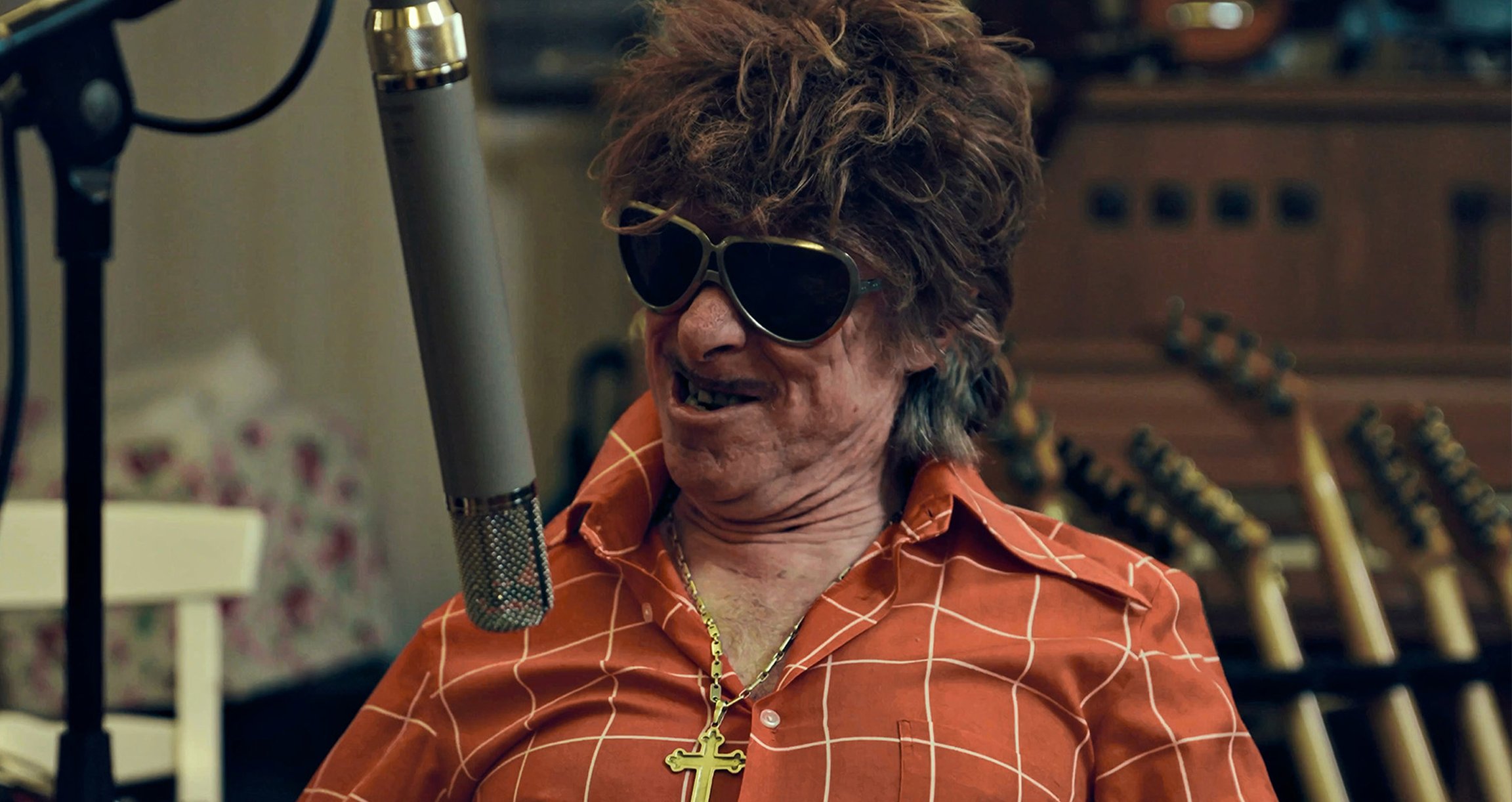 Helge Schneider grinst mit Sonnenbrille und einer zerzausten Frisur in einem orange karierten Hemd