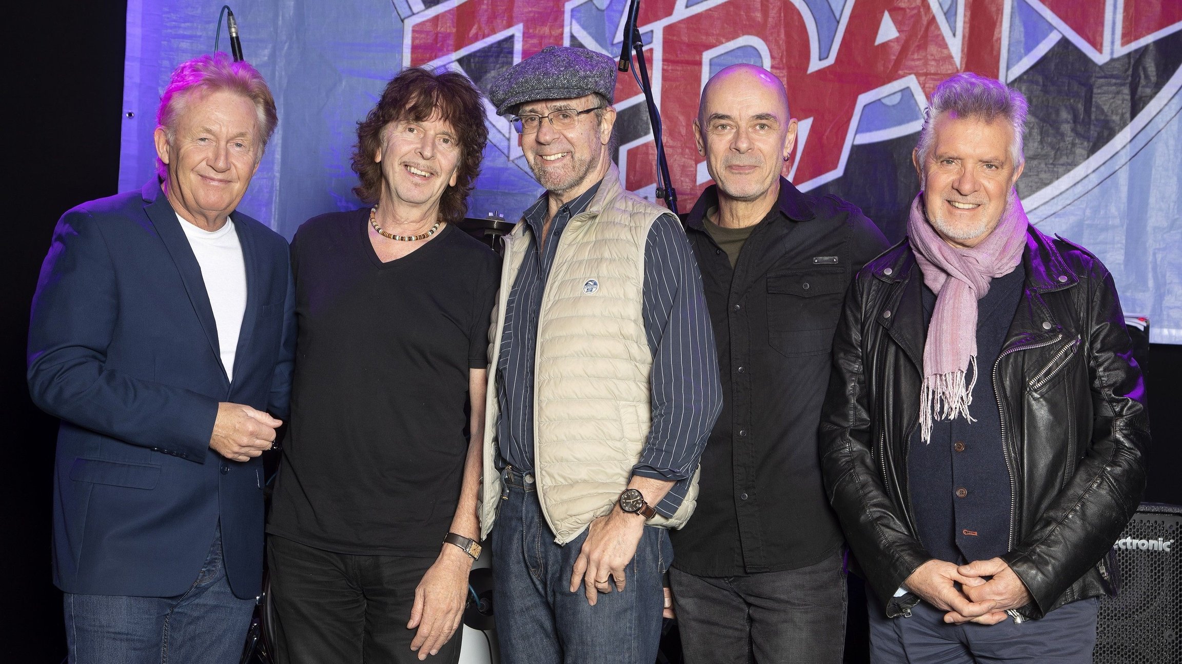 Die fünfköpfige Band Manfred Mann´s Earth Band in Freizeitkleidung steht vor einem Banner mit roten Buchstaben auf violettem Untergrund