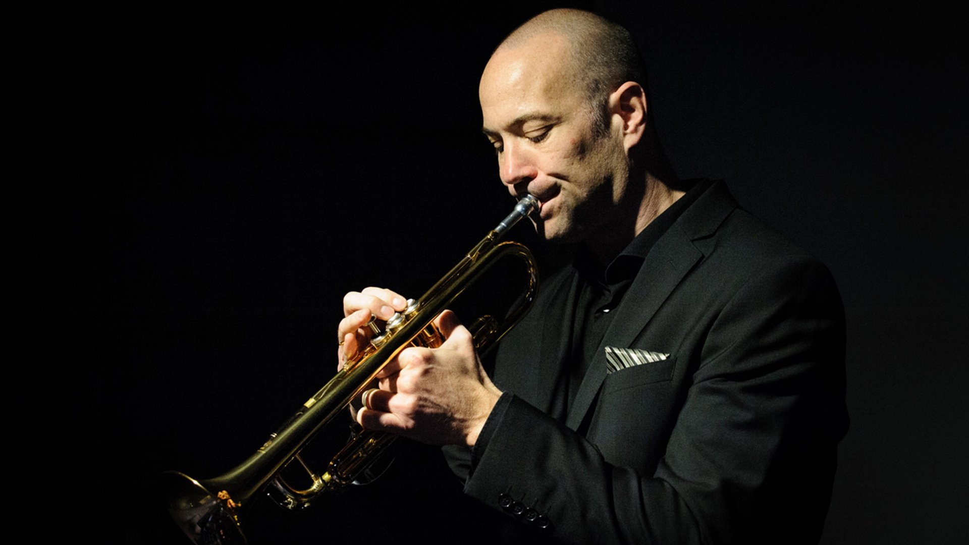 Auftritt von Florian Brandl (Trompete): Eine Person spielt eine goldfarbene Trompete in einem dunklen Bühnen‑Setting; die Szene ist gezielt ausgeleuchtet, der Fokus liegt auf Instrument, Händen und Oberkörper vor schwarzem Hintergrund, Studio‑ bzw. Konzertaufnahme.
