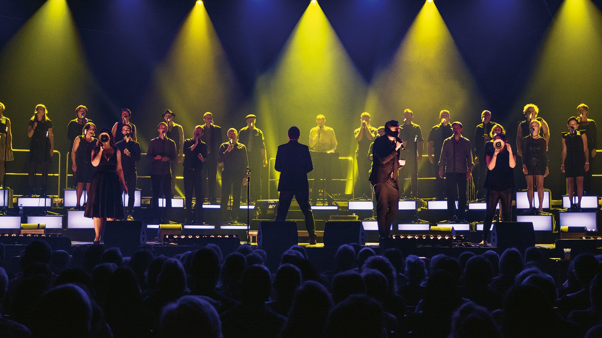 Zahlreiche Sänger auf einer Bühne, im Vordergrund das Publikum