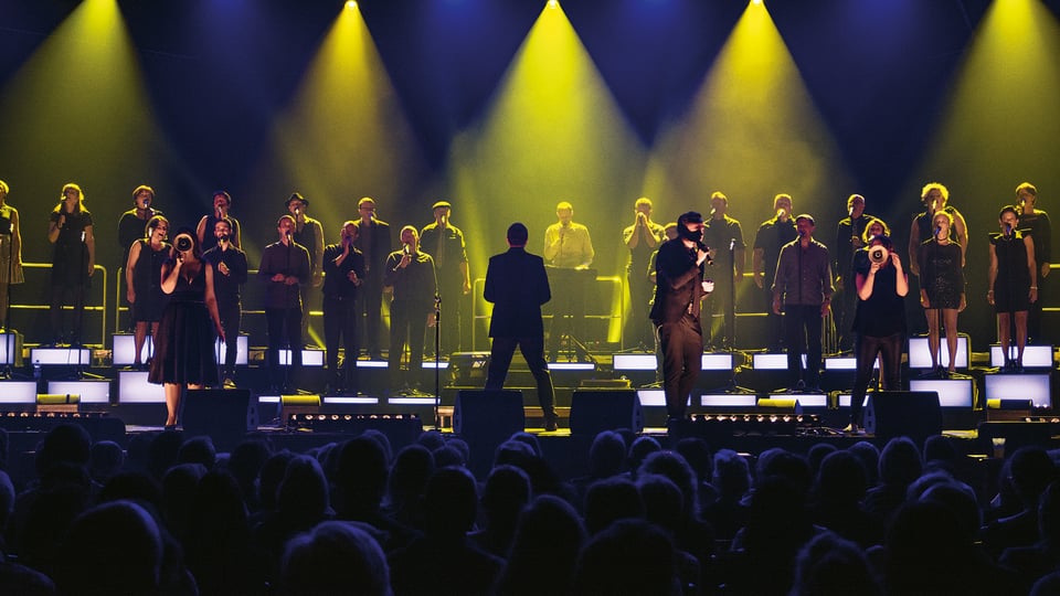 Zahlreiche Sänger auf einer Bühne, im Vordergrund das Publikum