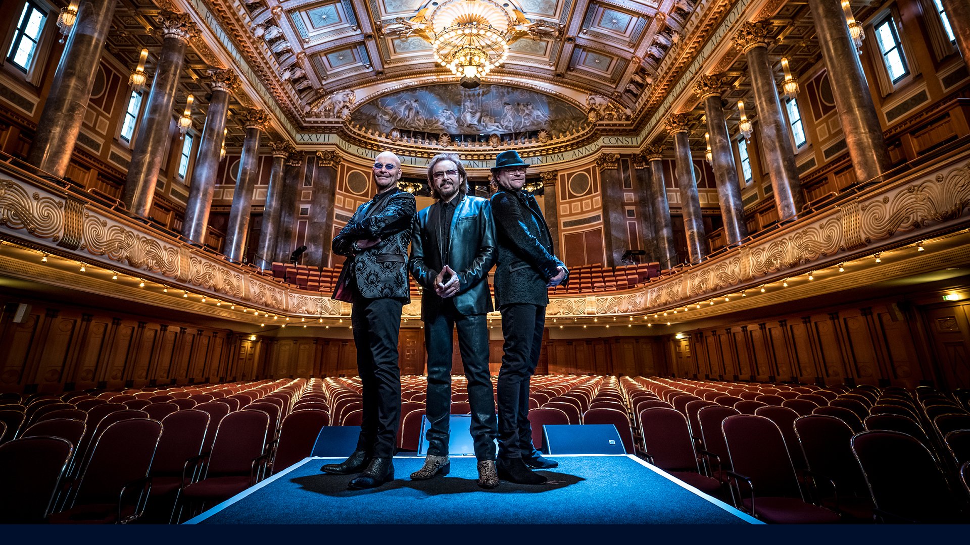 Motiv Nights on Broadway mit drei Personen in dunklen Anzügen die auf einer Bühne in einem prachtvollen Konzertsaal mit Kronleuchter und rot gepolsterten Sitzreihen stehen
