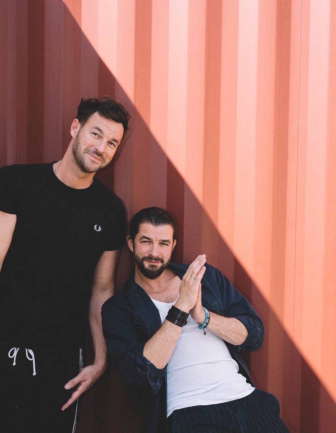 Die beiden Künstler schauen freundlich in die Kamera, sie befinden sich vor einem hellen Hintergrund.