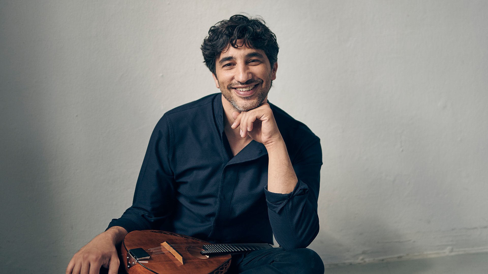 Avi Avital sitzt mit einem Saiteninstrument auf dem Schoß, lächelnd vor hellem Hintergrund