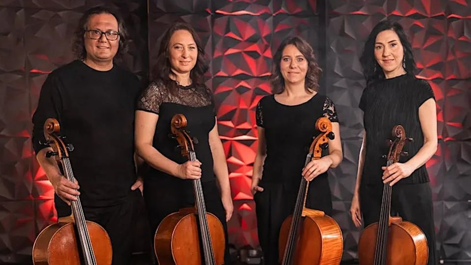 Vier Personen stehen nebeneinander vor strukturiertem, dunkel‑rotem Hintergrund und halten jeweils ein Cello.