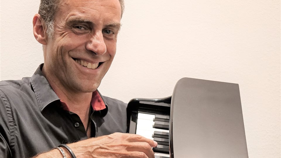 Foto für das Event „Martin Schmitt – Schmitt happens“ mit dem Künstler, der ein Klavier in den Händen hält, vor hellem Hintergrund.