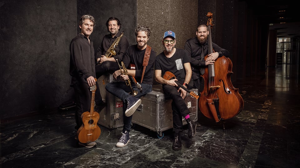 Die Band Söndörgő mit fünf Musikern, die gemeinsam vor einer dunklen Wand stehen und sitzen, einige halten Instrumente wie Saxophon, Kontrabass, Trompete und Saiteninstrumente; im Vordergrund stehen Instrumentenkoffer auf einem dunklen Boden.