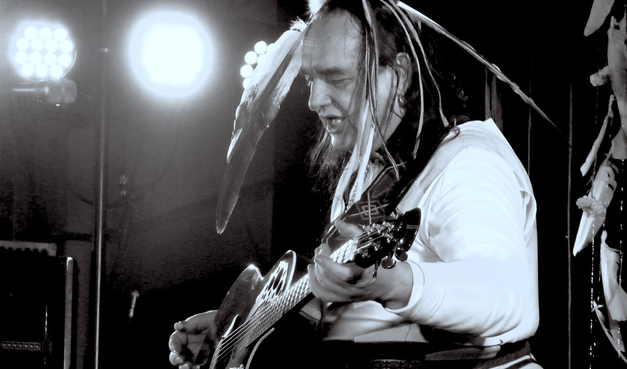 Schwarz-weiss Bild von Willy Michl mit Federschmuck auf dem Kopf und Gitarre auf einer Bühne