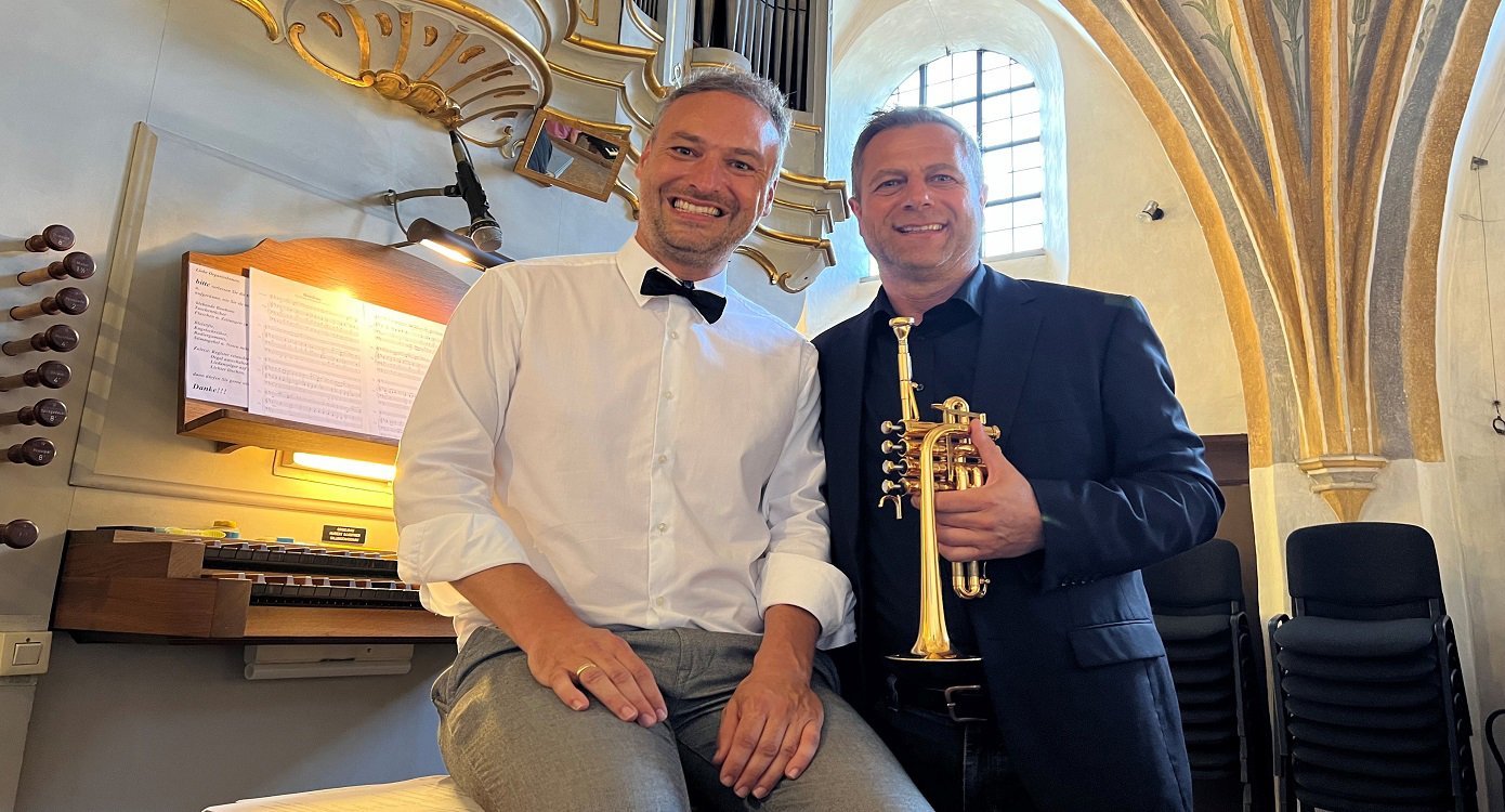 zwei Männer mit Instrumenten und einem Klavier im Hintergrund