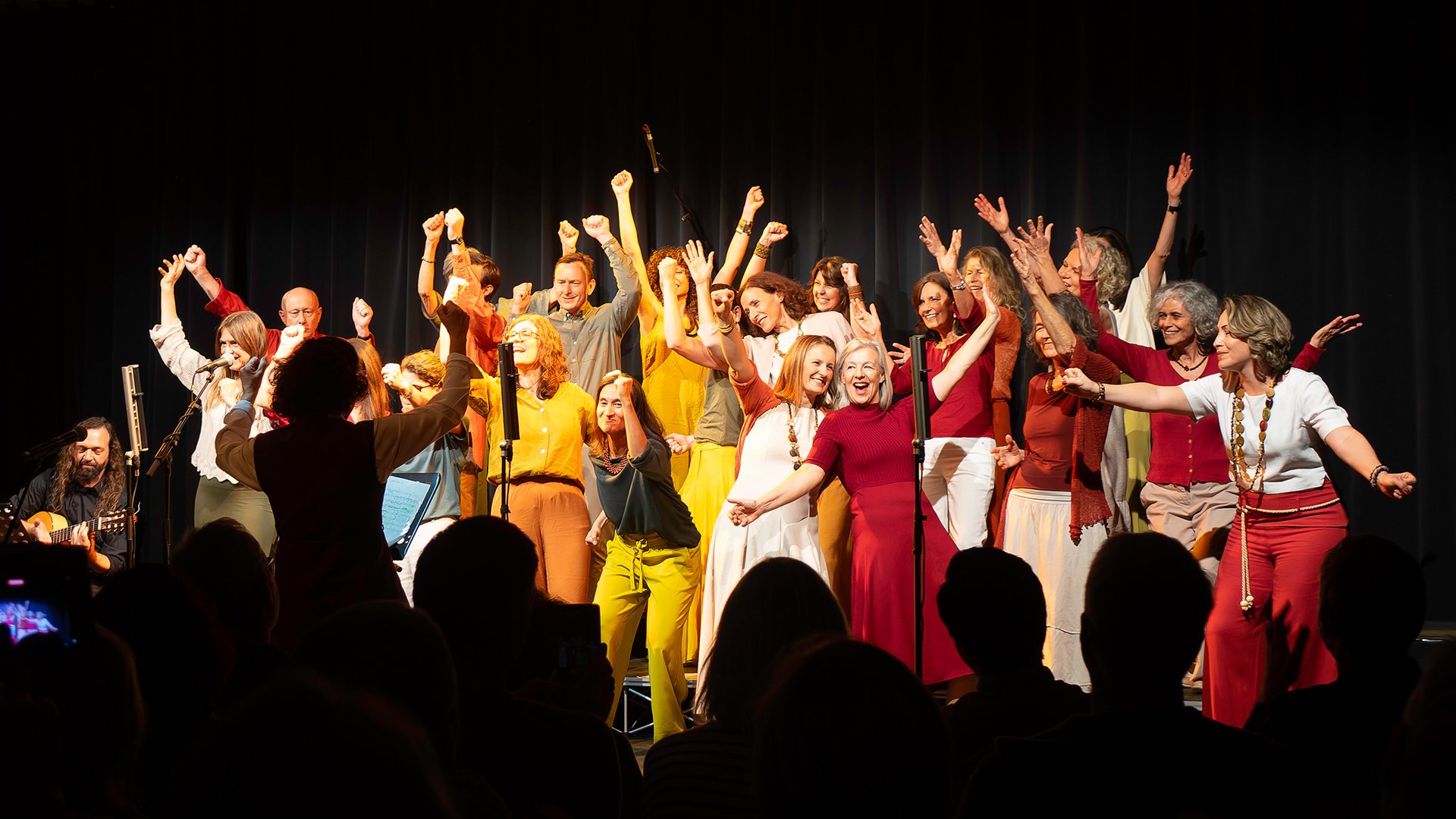 Große Gruppe von singenden Personen auf einer Bühne hebt die Arme; vorne dirigierende Person, im Vordergrund Publikum als Silhouetten