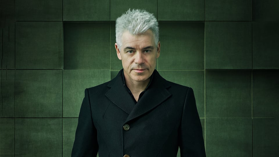 Der Künstler Michael Mittermeier in schwarzem Mantel vor einer Wand mit grünen quadratischen Kacheln